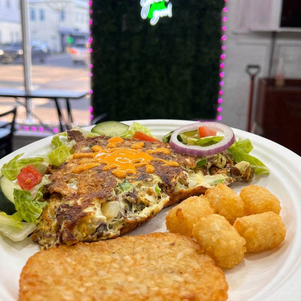 Plate of a cheesy, sauced omelet with lettuce, tomato, cucumber, and onion, served with tater tots in a casual restaurant.